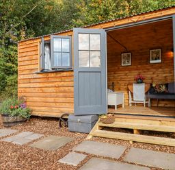 A cabin with open double doors and a window showing wooden interior with furniture and plants outside at Hideaway Cabin in Cadbury near Thorverton