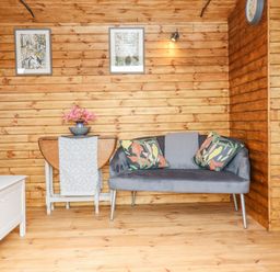 A wooden room with a gray sofa with patterned cushions a small table with flowers and two framed pictures on the wall at Hideaway Cabin in Cadbury near Thorverton