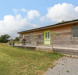 A wooden cabin with a green door and number 2 on the wall with chairs and plants on a grassy area at Lulu Logs in Congresbury