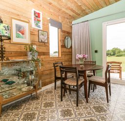 A dining area with a round wooden table and four chairs beside sliding glass doors and a glass cabinet against a wooden wall at Lulu Logs in Congresbury