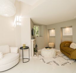 A living room with a white rounded armchair a small black side table a beige round ottoman a brown sofa and framed art on walls at The Hayloft in Sherston