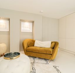 A living room with a mustard yellow sofa a white fur stool a round ottoman and built in wall shelves at The Hayloft in Sherston