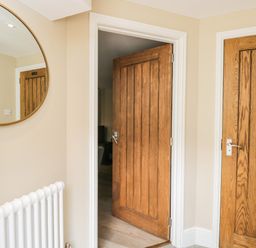 A hallway with two wooden doors one open and one labeled WC a round mirror and white radiator at The Coach House in Westonbirt near Tetbury