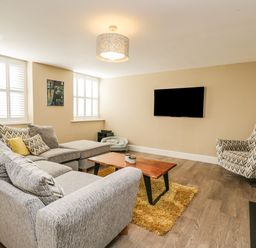 A living room with a grey sectional sofa a wooden coffee table a patterned armchair and a wall-mounted TV at The Coach House in Westonbirt near Tetbury
