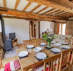 A dining area with a wooden table set with plates and glasses and a living area with a wood stove and a sofa at Munderfield Mill in Munderfield near Bishops Frome