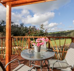 A covered wooden balcony with a glass table and two chairs overlooking fields and trees at Bearscombe View in Modbury