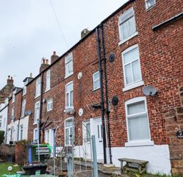 A row of brick terraced houses with white framed windows and satellite dishes with a garden and shed in front at Monteva in Whitby