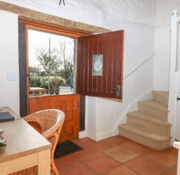 A hallway with an open wooden door a wicker chair a wooden table and carpeted stairs at The Old Barn in Colyton