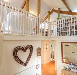 An interior view of a two-level room with wooden beams a white railing a large bookshelf and a heart-shaped wall decoration at The Old Barn in Colyton