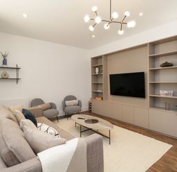 A living room with sofa, armchairs, coffee table, TV on a shelving unit, wall shelves, and plant at Alice House Stone Cross Mansion in Ulverston
