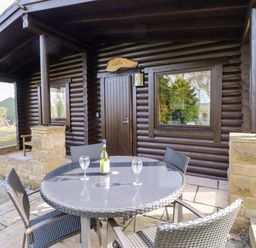 A patio with a round glass table, four chairs, a bottle of wine, and two wine glasses outside a log cabin at Alwinton Lodge in Longframlington