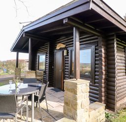 A log cabin with a covered porch and outdoor table with chairs outside at Alwinton Lodge in Longframlington