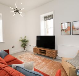 A living room with a red sofa blue and gray cushions a beige armchair a wooden TV stand a flat screen TV two abstract paintings a plant and two windows at William Tower Suite Stone Cross Mansion in Ulverston