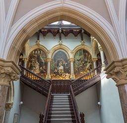 A grand staircase with wooden railings and large arches framing murals on the wall at The Alfred Suite Stone Cross Mansion in Ulverston