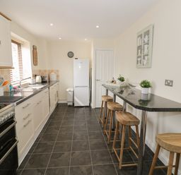 A kitchen with white cabinets black countertops stools along a bar and a white refrigerator at The Wheat House in Welford-On-Avon