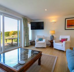 A living room with two white armchairs a blue sofa a wooden coffee table and sliding glass doors at Alpha in St Minver
