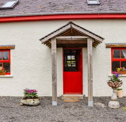 The exterior front of a house with a red door and two windows with red frames and flower pots at Cob Cottage in Kilmore County Wexford