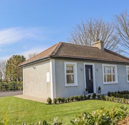A single-story house with three windows and a central door surrounded by grass and small bushes at An Teachain in Adamstown County Wexford