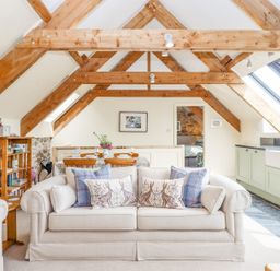 An open living space with wooden beams a beige sofa with cushions a dining table and a kitchen area with light green cabinets at The Coach House at Thorn Farm Bridford near Moretonhampstead