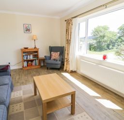 A living room with a wooden coffee table a blue sofa a blue armchair beside a bookshelf and a large window showing trees at Ceris Penrhyndeudraeth