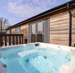An outdoor hot tub next to a wooden building with glass doors and a wooden railing at Skylarks in Fornham St Genevieve near Bury St Edmunds