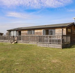 A wooden lodge with a fenced wooden deck on a grassy area at Skylarks in Fornham St Genevieve near Bury St Edmunds