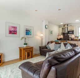 A living room with brown leather sofas a wooden TV stand a side table with nested stools and a kitchen area at Constantine Bay Cottage in Constantine Bay