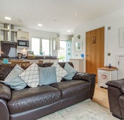 An open living room and kitchen with a brown leather sofa and armchair with cushions and kitchen appliances at Constantine Bay Cottage in Constantine Bay