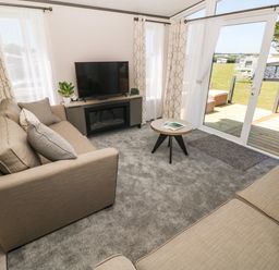A living room with two beige sofas a round coffee table a TV on a stand and sliding glass doors to a deck at Puffin Lodge in Broad Haven