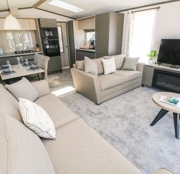 A living room with two beige sofas a flat screen tv on a cabinet a small round coffee table and a dining table and kitchen area at Puffin Lodge in Broad Haven