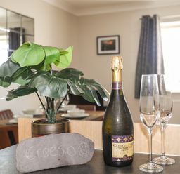 A kitchen counter with a potted plant a bottle of prosecco two empty champagne glasses and a stone with writing in a dining area at 4 Pen Llanw Tides Reach in Rhosneigr