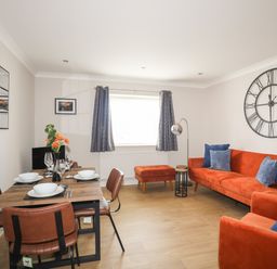A living and dining area with a wooden dining table set with dishes and glasses next to an orange sofa with blue cushions and a large wall clock at 4 Pen Llanw Tides Reach in Rhosneigr