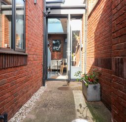 A narrow outdoor corridor with red brick walls on both sides a concrete planter with plants and an open glass door leading to a room with chairs at Farriers Retreat Budleigh Salterton