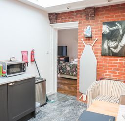 A kitchen area with a microwave on a counter ironing board on a brick wall and a striped chair at Farriers Retreat Budleigh Salterton