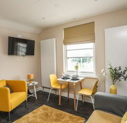 A living area with yellow armchairs a small dining table with yellow chairs a wall-mounted TV and a window with a brown blind at Cwm Gwyrfai in Caernarfon