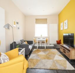 A living room with a yellow armchair a gray sofa a small dining table with yellow chairs a patterned rug and a wooden TV stand at Moel Tryfan in Caernarfon