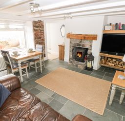 A living room with a brown leather sofa a dining table with two chairs a stone fireplace with a wood stove a television with firewood underneath and a small side table with a lamp at 1 Salt Cottage in Port Eynon