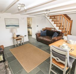 A living room with a brown leather sofa a wooden dining table with two place settings a small side table with a lamp and stairs in the background at 1 Salt Cottage in Port Eynon