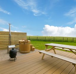 An outdoor deck area with a wooden picnic table a wooden hot tub and a black grill with a wooden fence and green fields at Green Fields in St Buryan