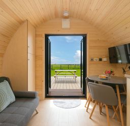 The interior of a wooden pod with a sofa pillow cabinet dining table chairs wall mounted tv and open doors to a deck with a picnic table at Green Fields in St Buryan