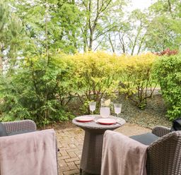 An outdoor patio area with two wicker chairs a small table with plates a jug and glasses surrounded by green bushes at Cosy Corner in the Lakes in Greystoke