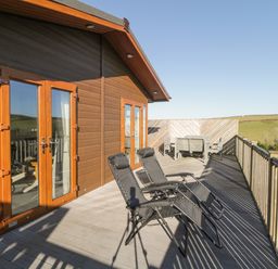 An outdoor wooden deck with two black lounge chairs and a table with chairs overlooking grassy hills at Ridges Retreat No 7 Kigbeare Lodges Ashbury Resort near Fowley Cross Sourton Down and Okehampton