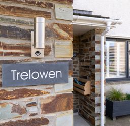 A stone wall with a sign reading Trelowen and a light fixture next to a window with white shutters and a planter box at Trelowen St Issey near Padstow