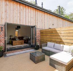 An outdoor patio area with gray cushioned wicker seating and a glass table next to a building with wooden and brick walls at Little Lugg in Cross Keys near Hereford