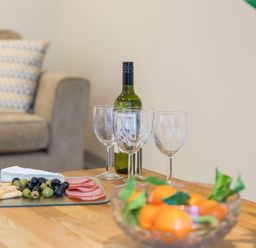 A table with wine glasses a bottle of wine a bowl of fruit and a platter of cheese olives and sliced meat in a living room with a couch and cushion at Golden Sands in Carbis Bay