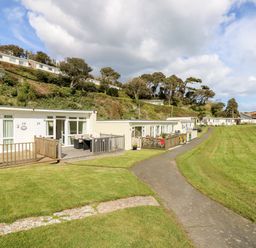 A row of single-story holiday homes with wooden decks along a paved pathway and a grassy hill with trees in the background at Seas The Day in Ventnor