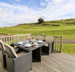 An outdoor dining area with a wicker table and chairs set on a wooden deck overlooking a grassy hill at Seas The Day in Ventnor