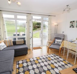 A living room with a gray sofa, an armchair, a dining table with chairs, a patterned rug, and large windows with curtains at Seas The Day in Ventnor