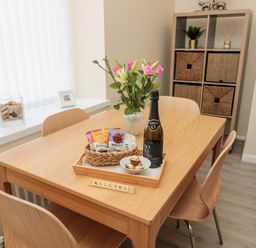 A dining room with a wooden table and chairs a vase with flowers a basket of snacks a bottle of wine and a storage unit with baskets at Stones Throw in Amble