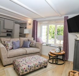 A sitting room with a beige sofa purple cushions floral patterned ottoman wooden nesting tables lamp and wall mounted tv at Hillside in Crafthole